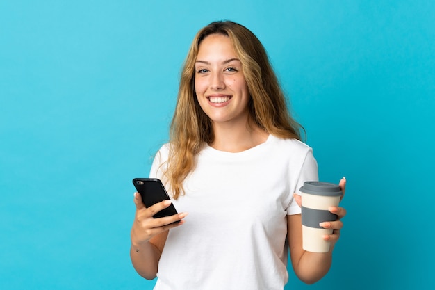 Young blonde woman isolated on blue wall holding coffee to take away and a mobile