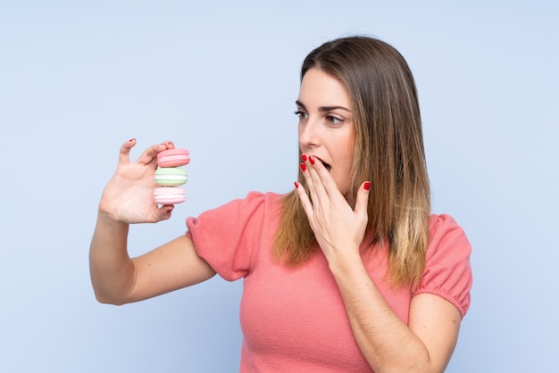 Young blonde woman over isolated blue wall holding colorful French macarons and with surprise expression