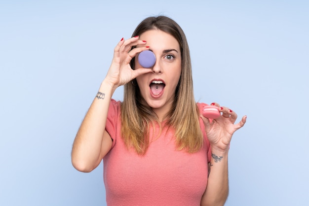 Young blonde woman over isolated blue wall holding colorful French macarons