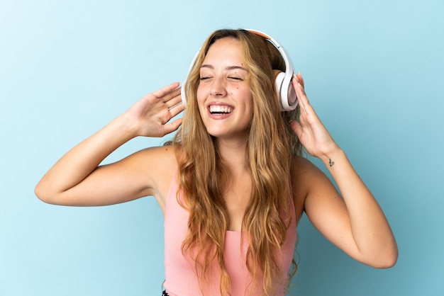 Young blonde woman isolated on blue wall listening music and singing