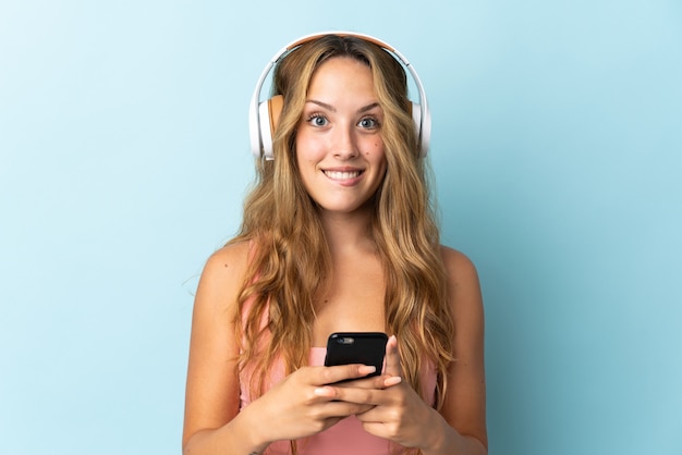 Young blonde woman isolated on blue wall listening music with a mobile and looking front