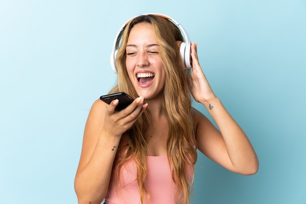 Young blonde woman isolated on blue wall listening music with a mobile and singing