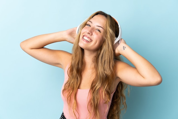 Young blonde woman isolated on blue wall listening music