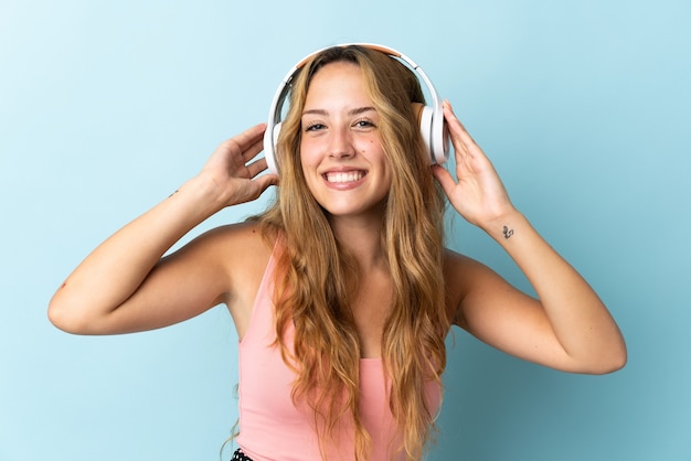 Young blonde woman isolated on blue wall listening music