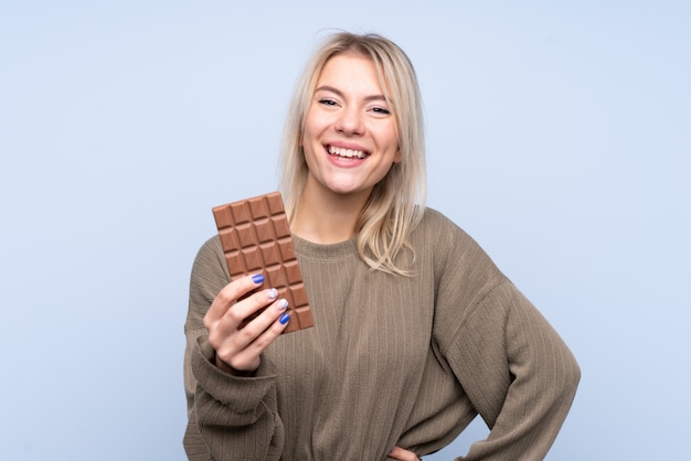 Young blonde woman over isolated blue wall taking a chocolate tablet and happy