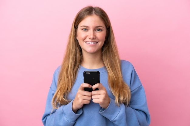 Young blonde woman isolated on pink background looking at the camera and smiling while using the mobile