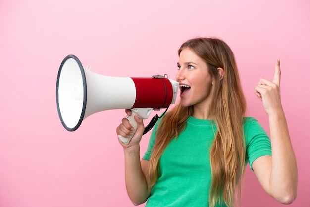 Young blonde woman isolated on pink background shouting through a megaphone to announce something in lateral position