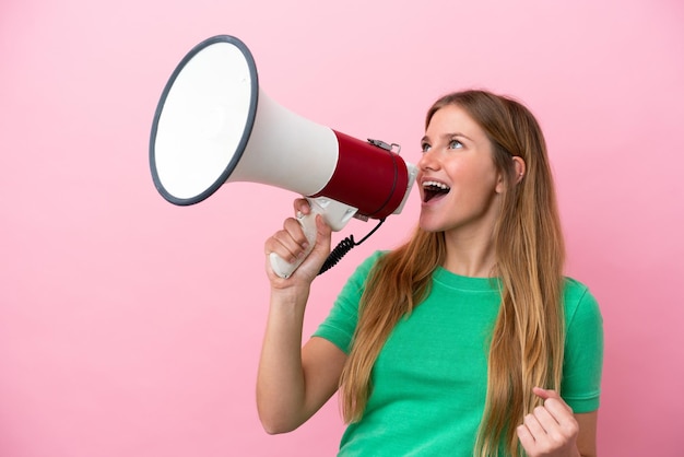 Young blonde woman isolated on pink background shouting through a megaphone to announce something in lateral position