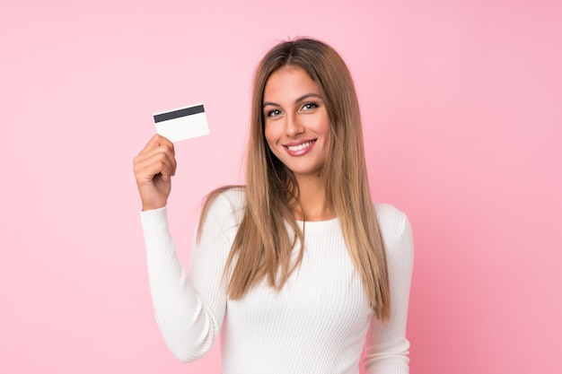 Young blonde woman over isolated pink wall holding a credit card