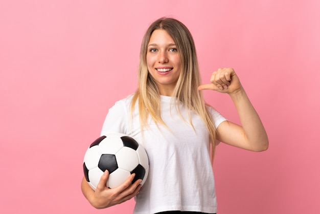 Young blonde woman isolated on pink wall with soccer ball and proud of herself