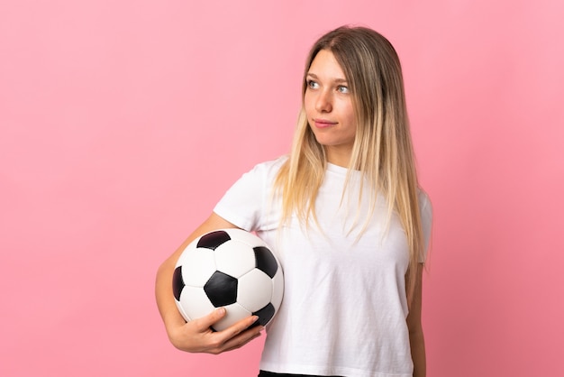 Young blonde woman isolated on pink wall with soccer ball