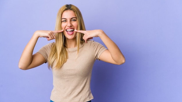 Young blonde woman isolated on purple smiles, pointing fingers at mouth.