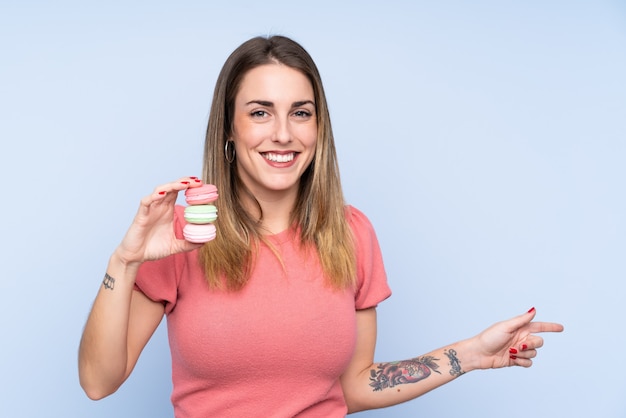 Young blonde woman over isolated wall holding colorful French macarons and pointing side