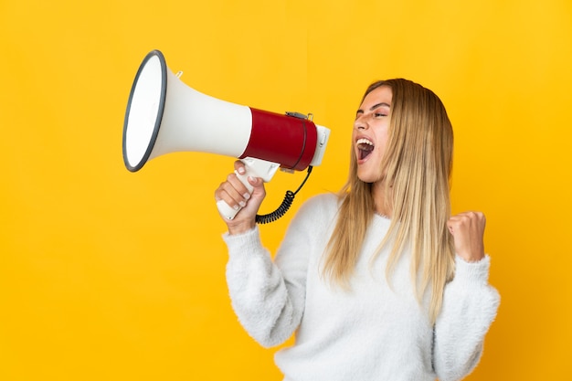 Young blonde woman over isolated wall shouting through a megaphone to announce something in lateral position