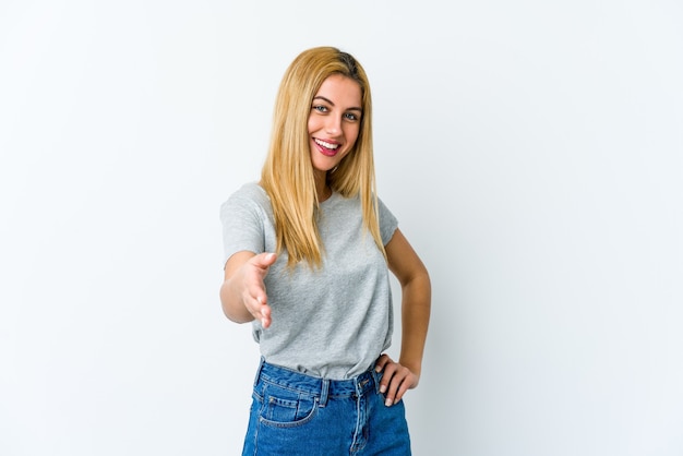 Young blonde woman isolated on white background stretching hand at camera in greeting gesture.