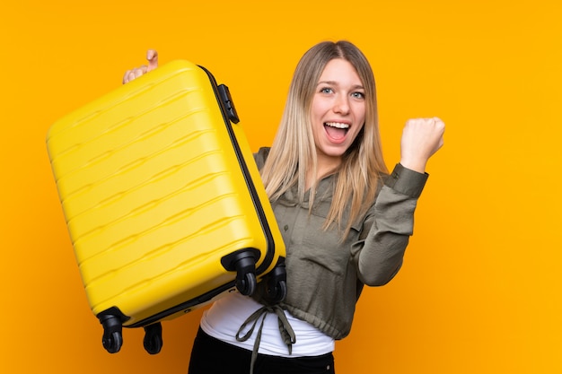 Young blonde woman over isolated yellow wall in vacation with travel suitcase