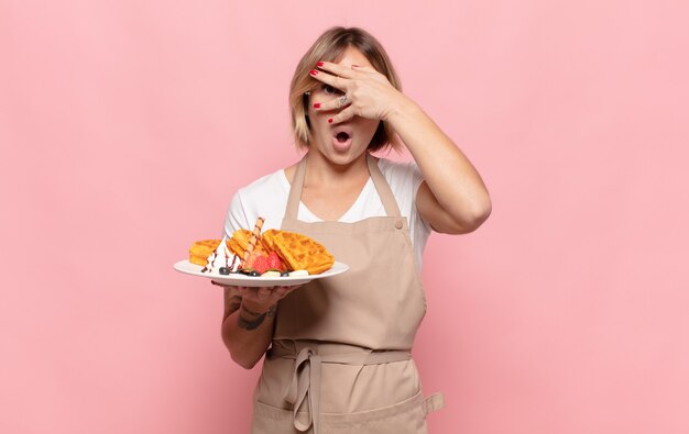 Young blonde woman looking shocked, scared or terrified, covering face with hand and peeking between fingers