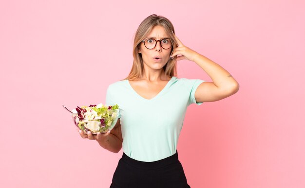 Young blonde woman looking surprised, realizing a new thought, idea or concept and holding a salad