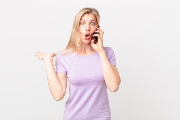 Young blonde woman looking surprised and shocked, with jaw dropped holding an object and calling with a smart phone