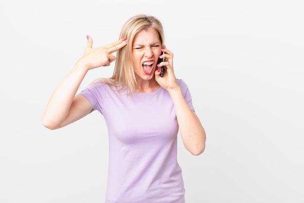 Young blonde woman looking unhappy and stressed, suicide gesture making gun sign and calling with a smart phone