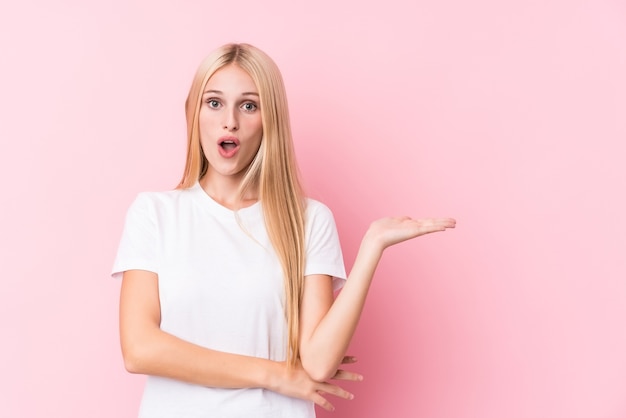 Young blonde woman on pink background impressed holding copy space on palm.