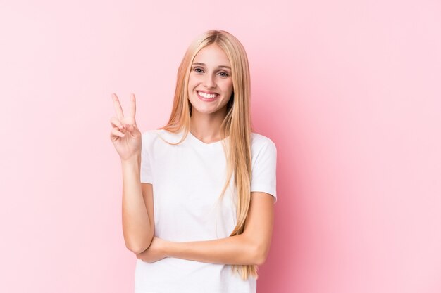 Young blonde woman on pink wall showing number two with fingers.