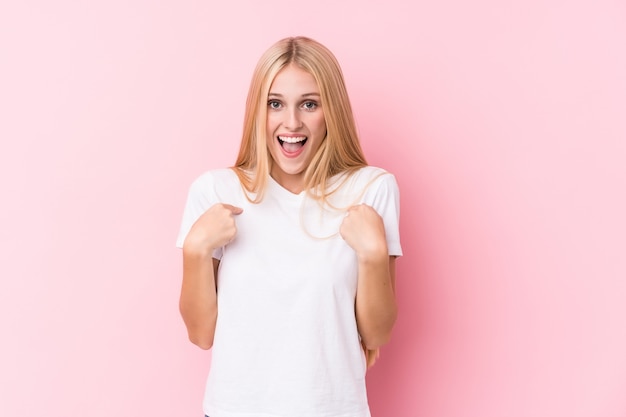 Young blonde woman on pink wall surprised pointing with finger, smiling broadly.