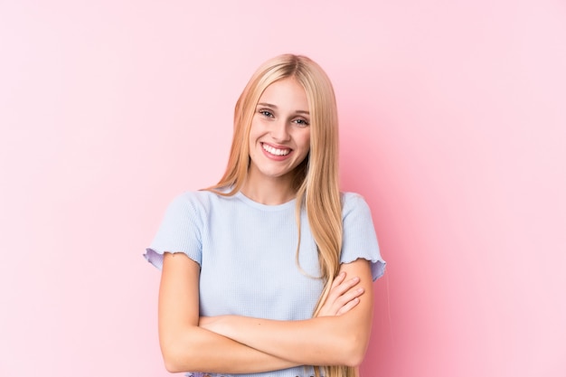 Young blonde woman on pink wall who feels confident, crossing arms with determination.