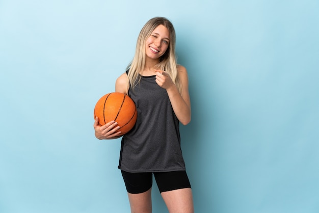 Young blonde woman playing basketball isolated on blue wall points finger at you