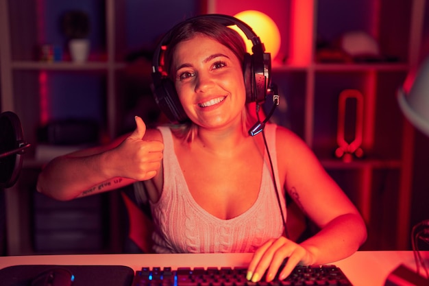 Young blonde woman playing video games wearing headphones doing happy thumbs up gesture with hand. approving expression looking at the camera showing success.