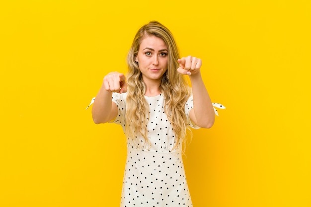 Young blonde woman pointing forward at camera with both fingers and angry expression, telling you to do your duty against yellow wall