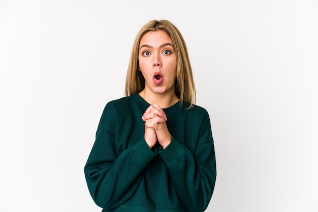 Young blonde woman praying for luck, amazed and opening mouth looking to front