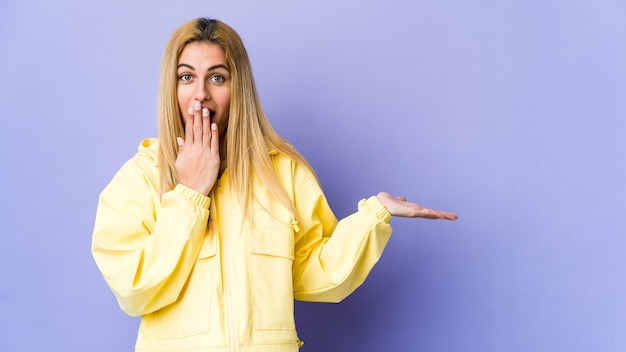 Young blonde woman on purple wall holding product on palm.