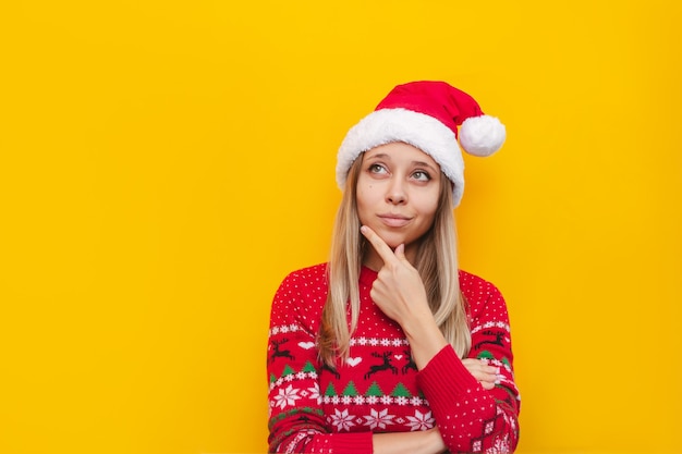 A young blonde woman in a red deer  sweater and a Santa Claus hat thinks looking away at copy space