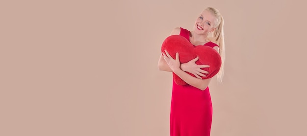 Young blonde woman in a red dress holds a red heart in her hands on a beige background