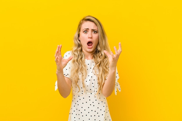 Young blonde woman screaming with hands up in the air, feeling furious, frustrated, stressed and upset against yellow wall