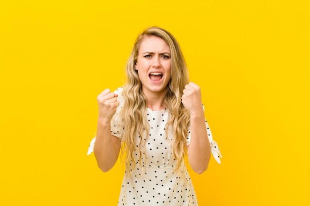 Young blonde woman shouting aggressively with an angry expression or with fists clenched celebrating success against yellow wall