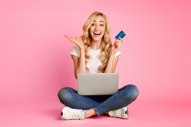 Photo a young blonde woman sits crosslegged on a pink background smiling excitedly as she holds a credit