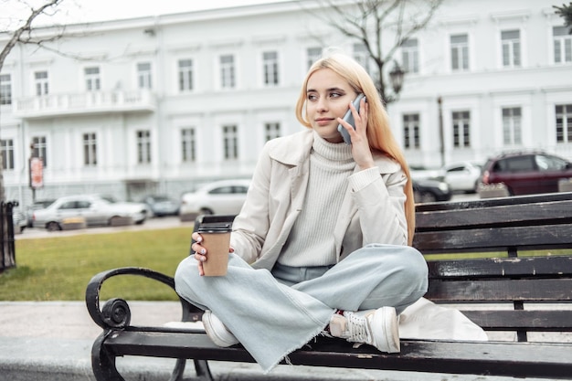 Young blonde woman sitting on a bench talking on the phone and drinking coffee