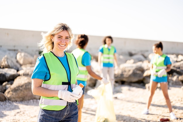 Young blonde woman smiling at the camera while her teamwork cleaning the rubish on a beach volunteers collecting the waste on the coast line young people working in team aware of the pollution