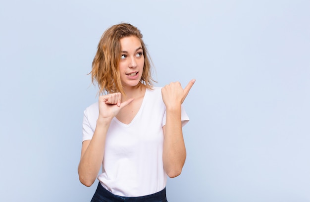 Young blonde woman smiling cheerfully and casually pointing to copy space on the side, feeling happy and satisfied against flat color wall