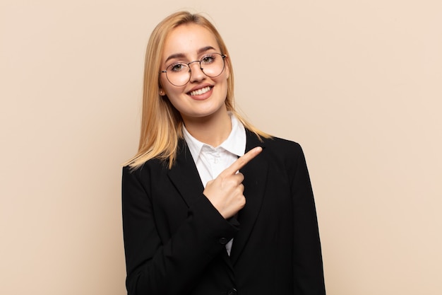 Photo young blonde woman smiling cheerfully, feeling happy and pointing to the side and upwards, showing object in copy space