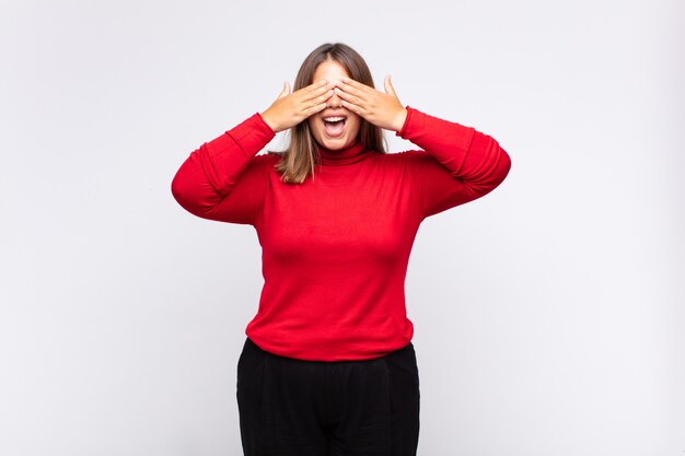 Young blonde woman smiling and feeling happy, covering eyes with both hands and waiting for unbelievable surprise