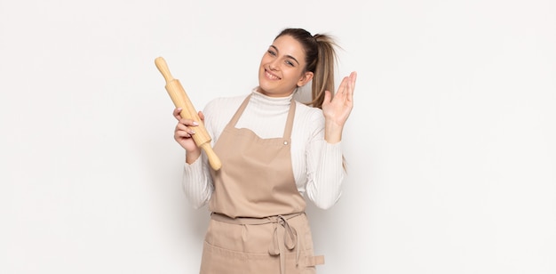 Young blonde woman smiling happily and cheerfully, waving hand, welcoming and greeting you, or saying goodbye