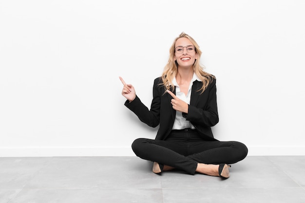 Young blonde woman smiling happily and pointing to side and upwards with both hands showing object in copy space sitting on the floor