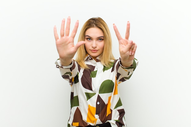 Photo young blonde woman smiling and looking friendly, showing number seven or seventh with hand forward, counting down over white wall