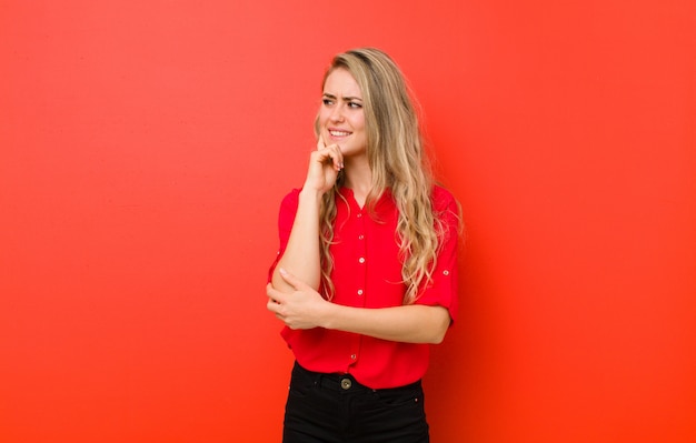 Young blonde woman smiling with a happy, confident expression with hand on chin, wondering and looking to the side against red wall