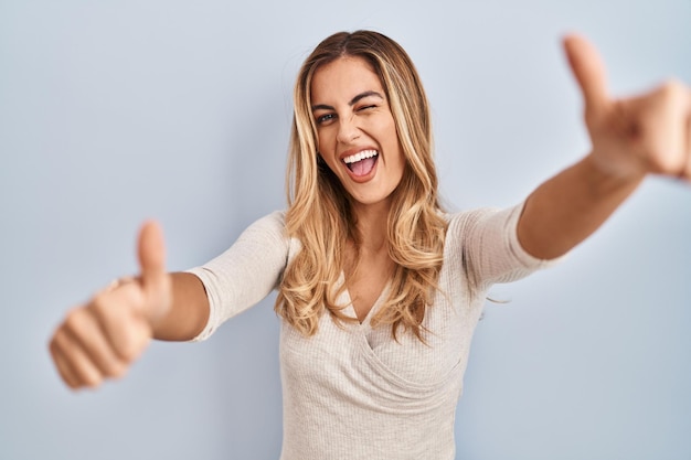 Young blonde woman standing over isolated background approving doing positive gesture with hand thumbs up smiling and happy for success winner gesture