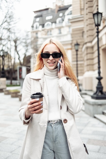 Young blonde woman in sunglasses talking on the phone and holding a cup of coffee in city