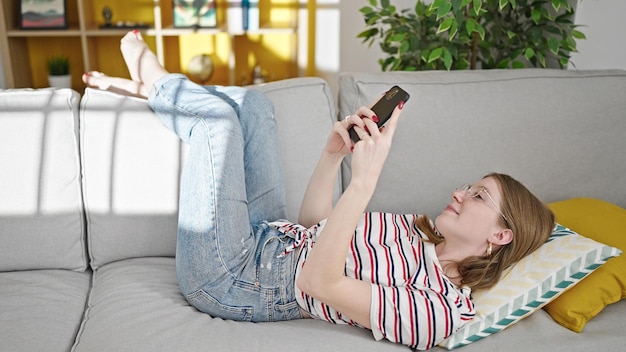 Young blonde woman using smartphone lying on sofa at home